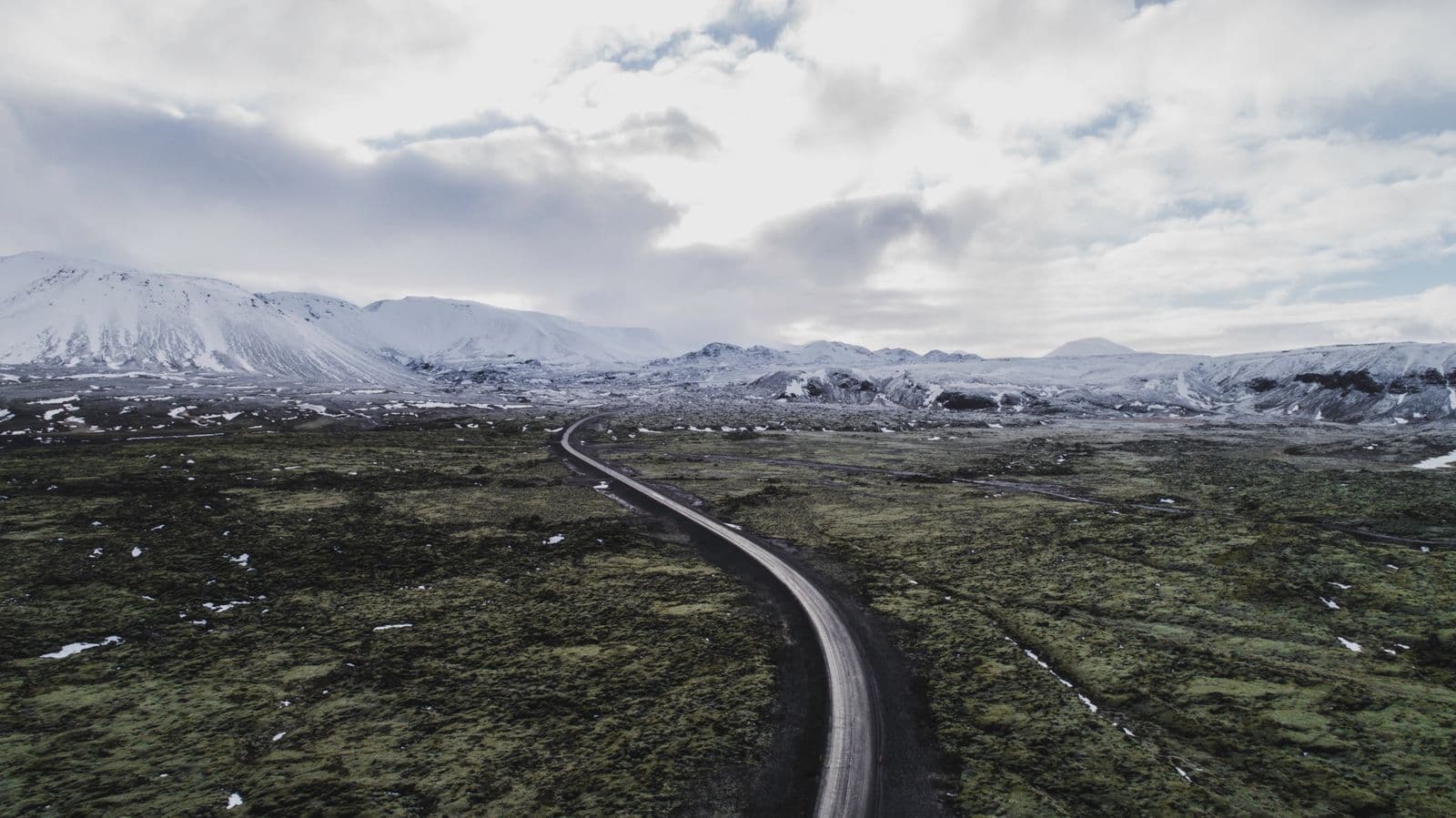 A lone highland road cutting through mossy lava fields toward snow-covered mountains