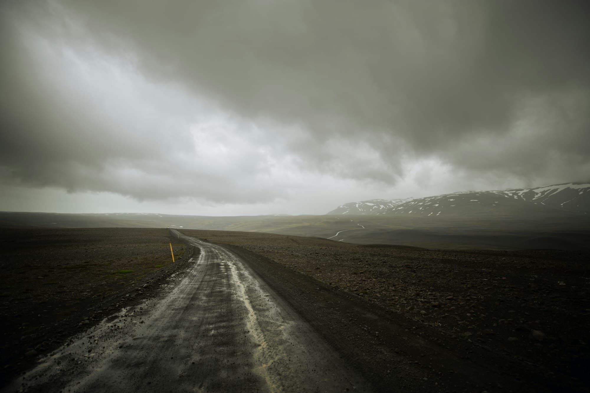 Leere Straße, die sich unter einem dramatischen grauen Himmel durch Islands grüne Hochlandlandschaft zieht